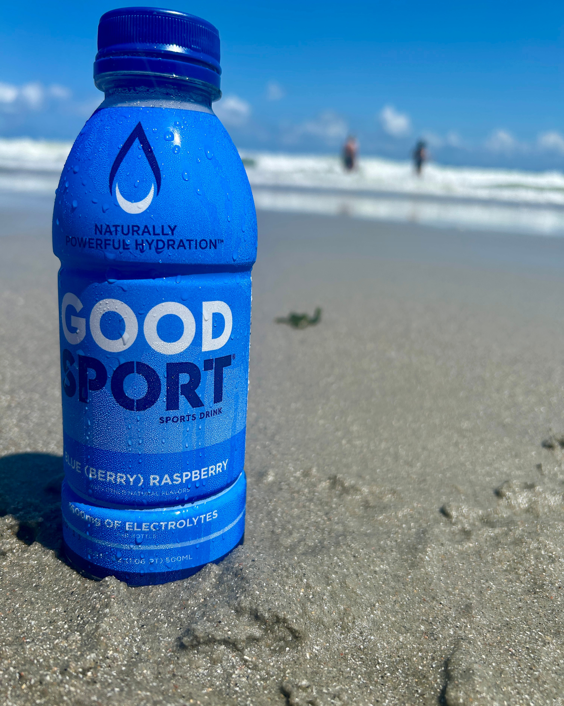 GoodSport sports drink Blue(berry) Raspberry bottle on a sandy beach with ocean waves in the background