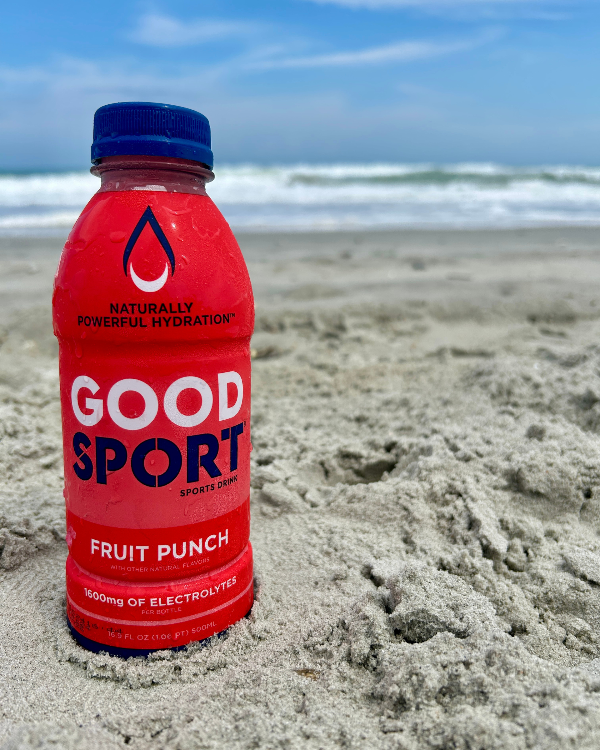Bottle of GoodSport Fruit Punch bottle on the sand a the beach. Blue skies and waves crashing in the background.